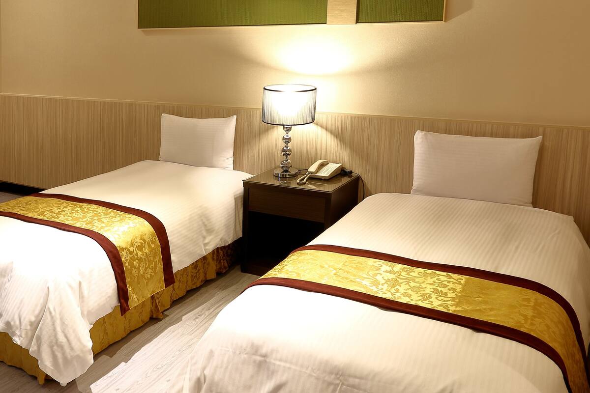 A hotel room with two single beds, each with a white and gold bedspread, a bedside table with a lamp, and a telephone.