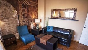 A living room with a stone wall, a black leather sofa, a blue armchair, a wooden coffee table, and a small side table with a lamp. - Studio Orient (Belgrade)
