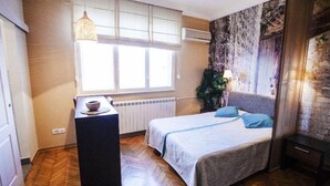 A bedroom with a bed, a small table, a window with blinds, a radiator, and a wall with a textured mural. - Studio Orient (Belgrade)