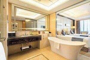 Un baño de hotel moderno con un espejo grande, una bañera independiente, un tocador con lavamanos y una cama con vistas a la ciudad.