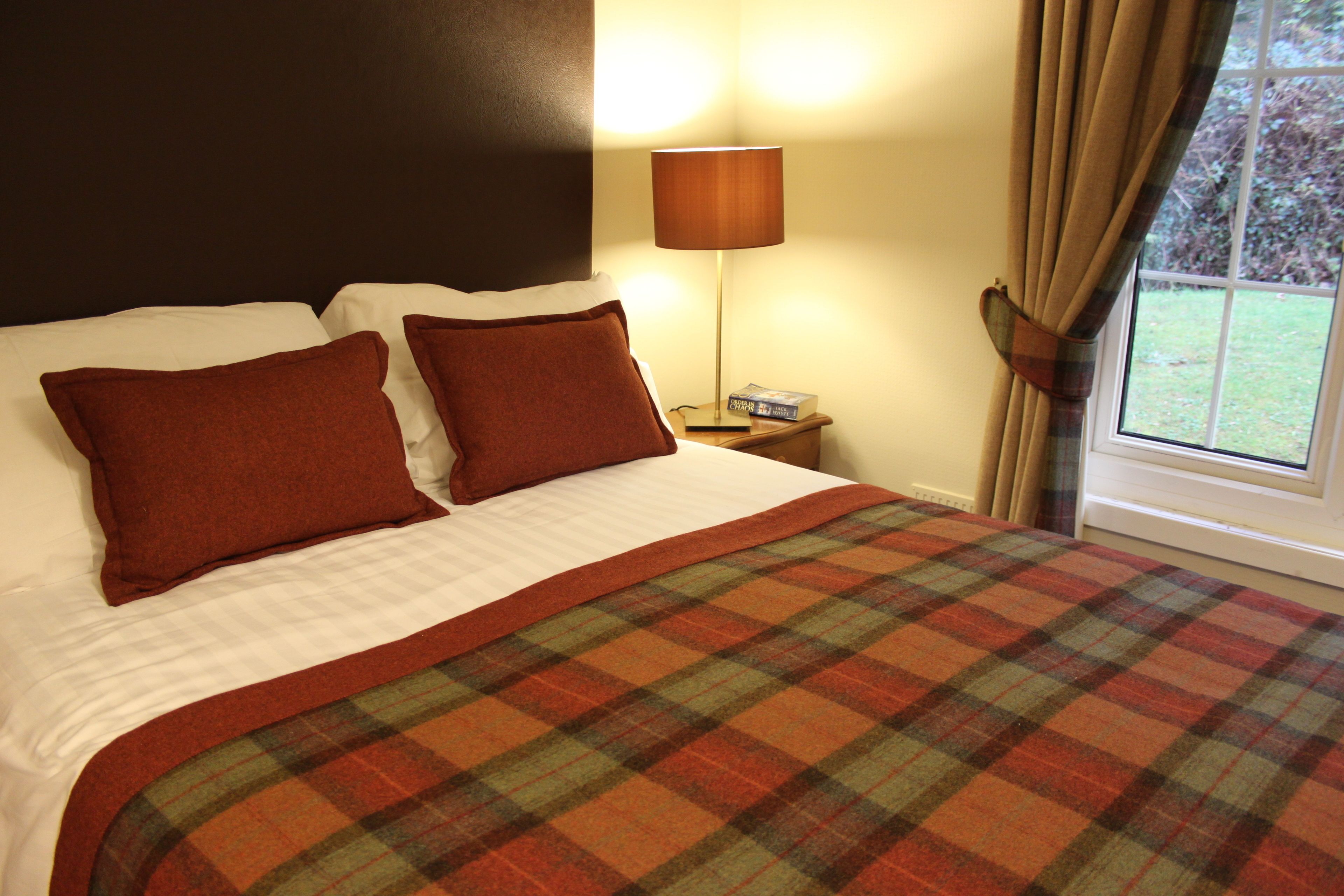 A neatly made bed with a plaid blanket, two brown pillows, a bedside lamp, and a window with curtains.