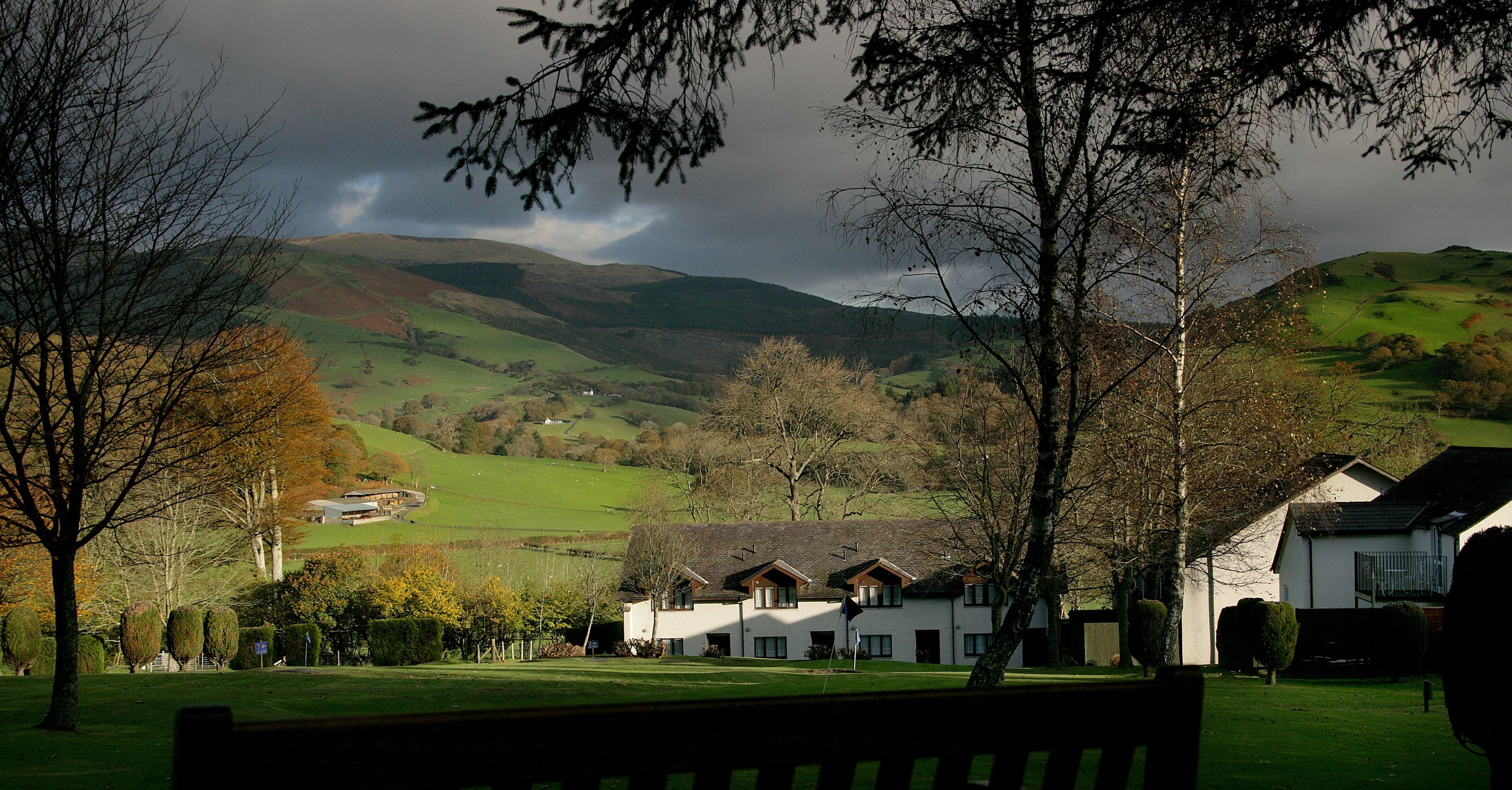A rural landscape with rolling hills, a white building with multiple windows, and a bench in the foreground.