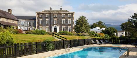 A swimming pool with a stone border, a black fence, and a large building in the background.