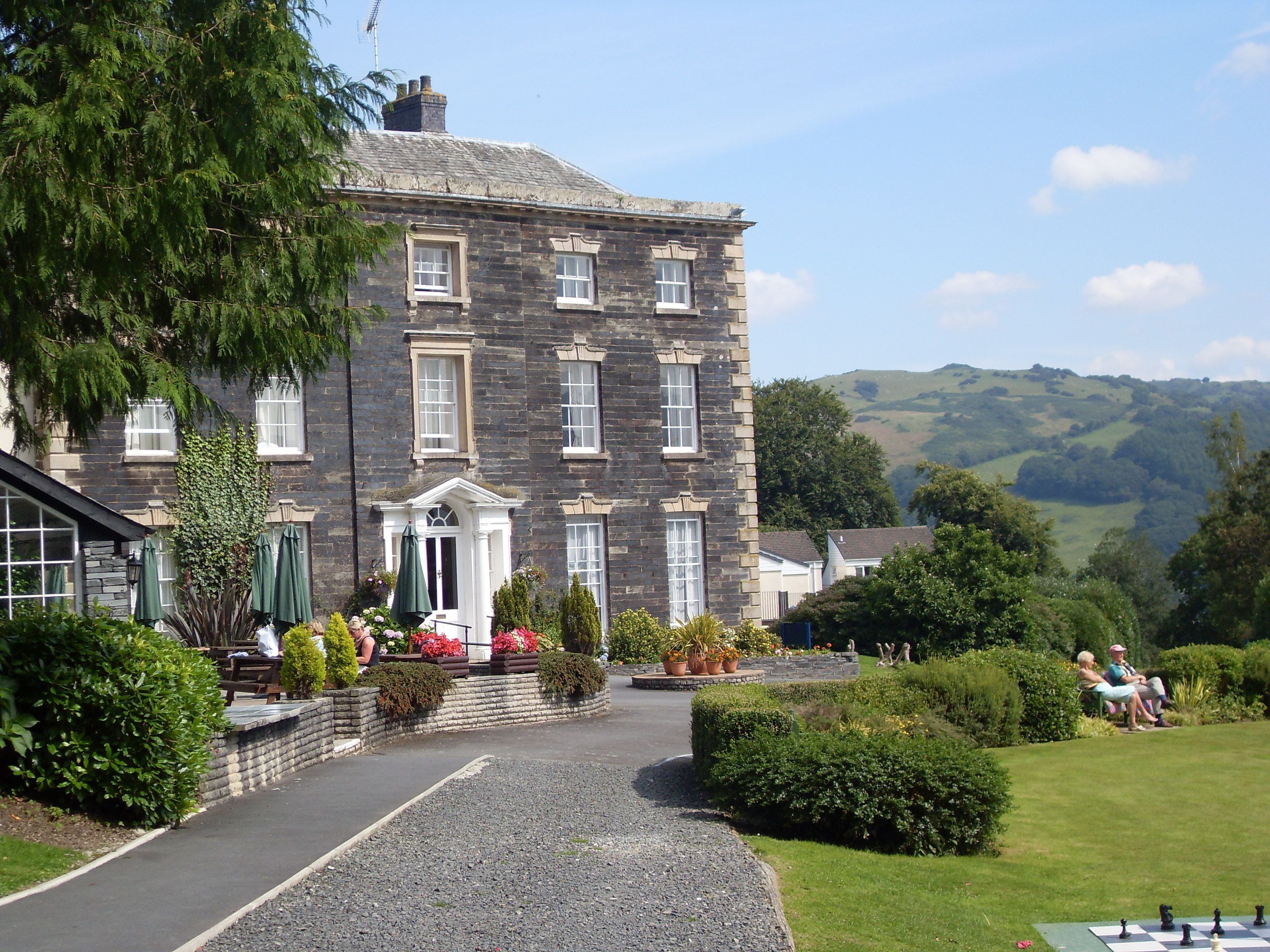 A stone building with a central entrance, surrounded by well-maintained gardens and a gravel pathway leading to it.