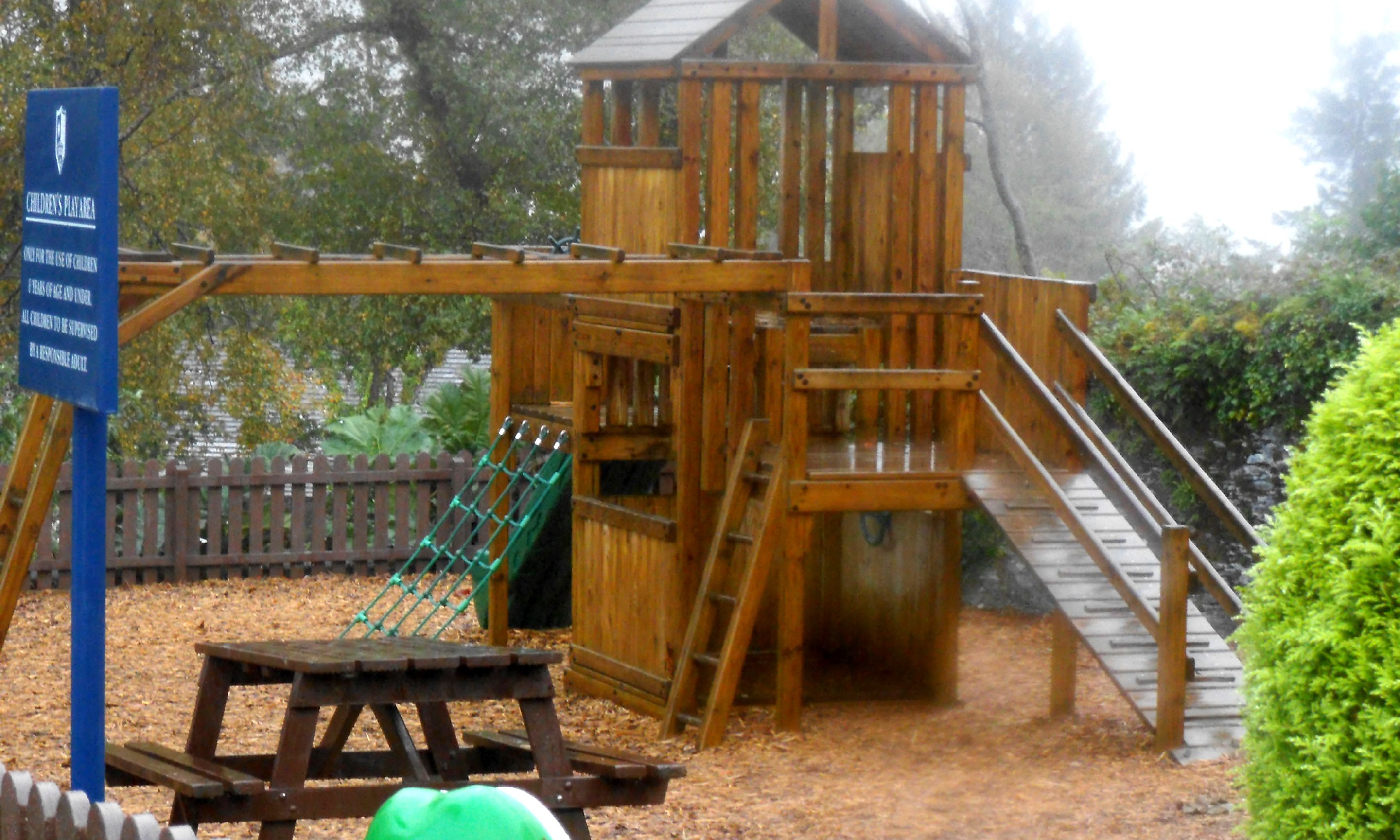 A wooden children's play area with a slide, climbing structures, and a picnic table.