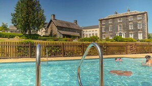 A swimming pool with a wooden fence and metal ladder, surrounded by lush greenery and historic buildings.