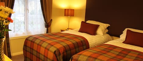 Quarto de hotel com duas camas, mesa de cabeceira, abajur e janela com cortinas.