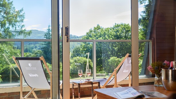 A room with two beach chairs, a table with a glass of wine, and a view of a forested area through large windows.
