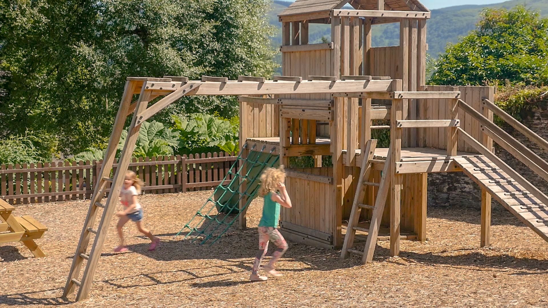 A wooden playground structure with slides and climbing elements, surrounded by a wooden fence and trees.