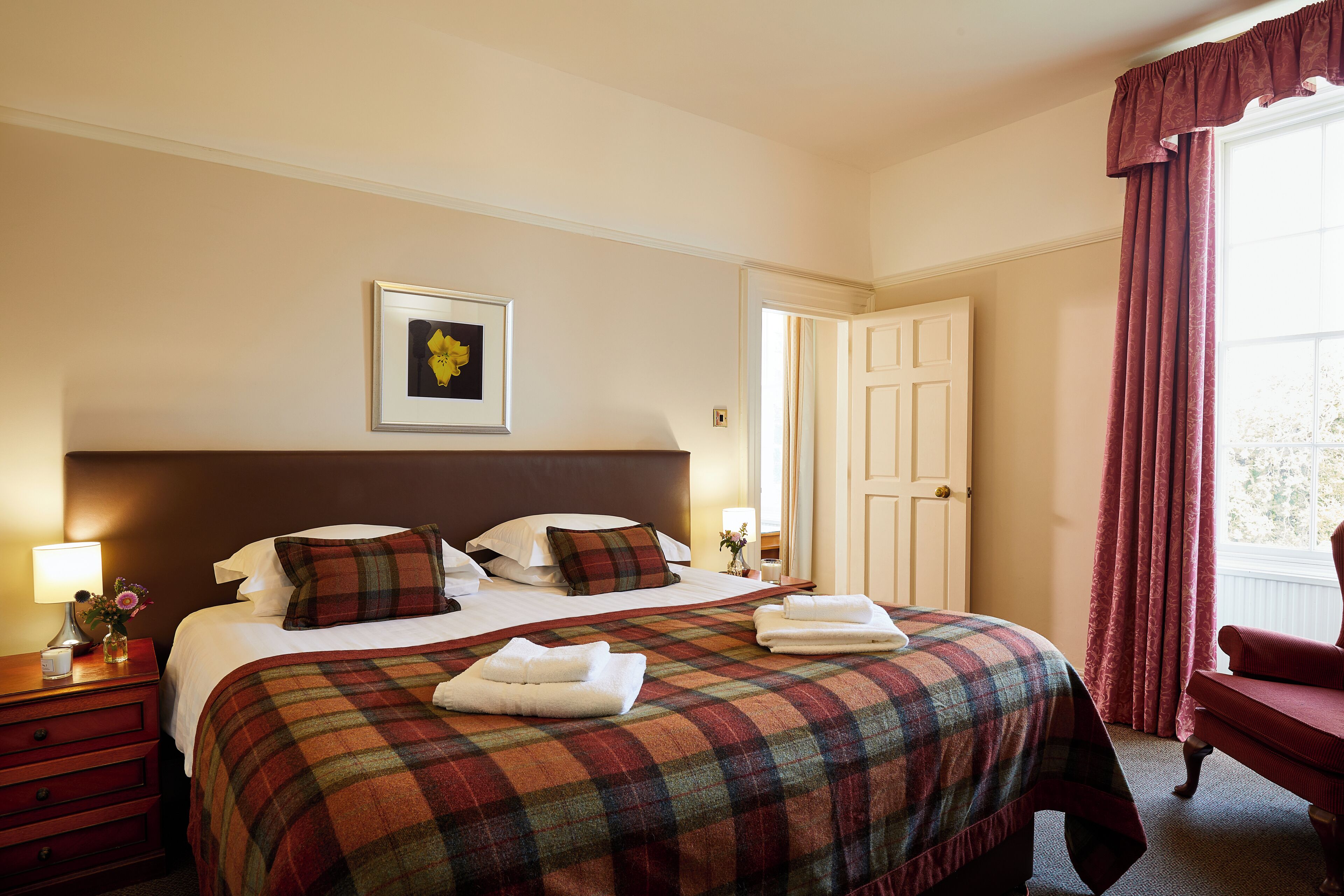Una habitación de hotel con una cama grande, una mesita de noche con lámpara, un cuadro en la pared y una puerta que da a otra habitación.