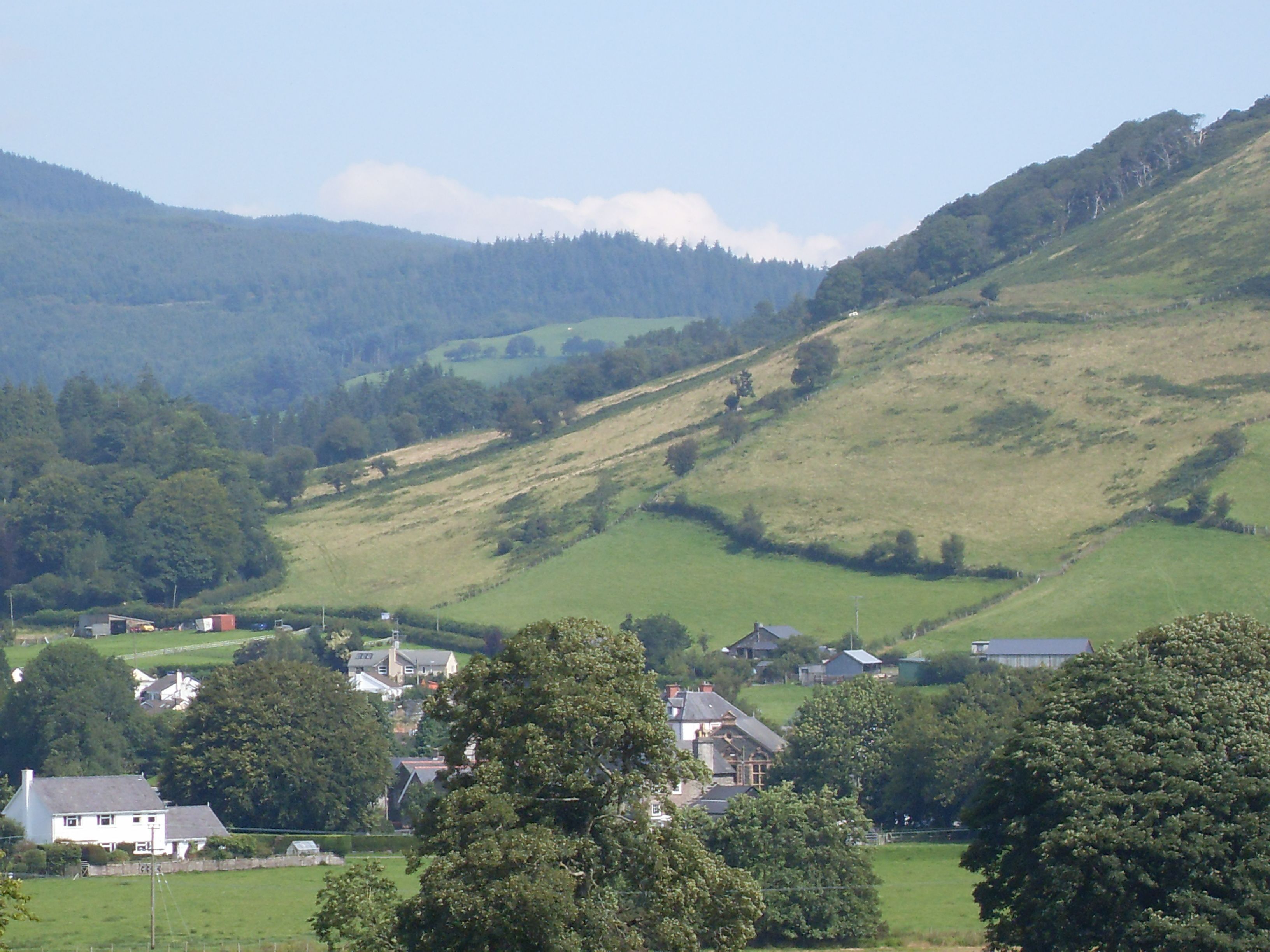 A rural landscape with rolling hills, scattered houses, and a mix of open fields and wooded areas.