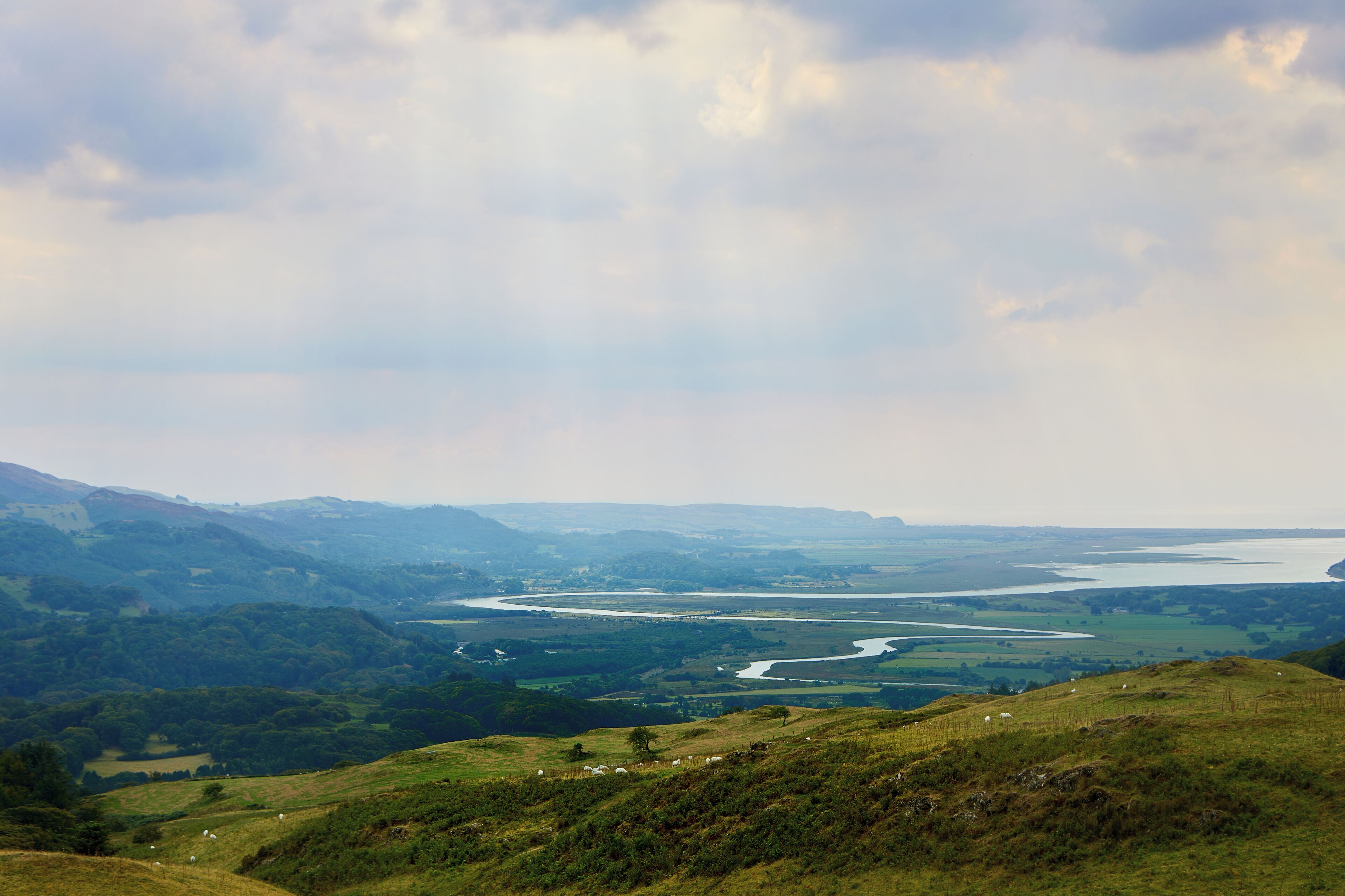 A panoramic view of a river winding through a valley with hills and a cloudy sky.
