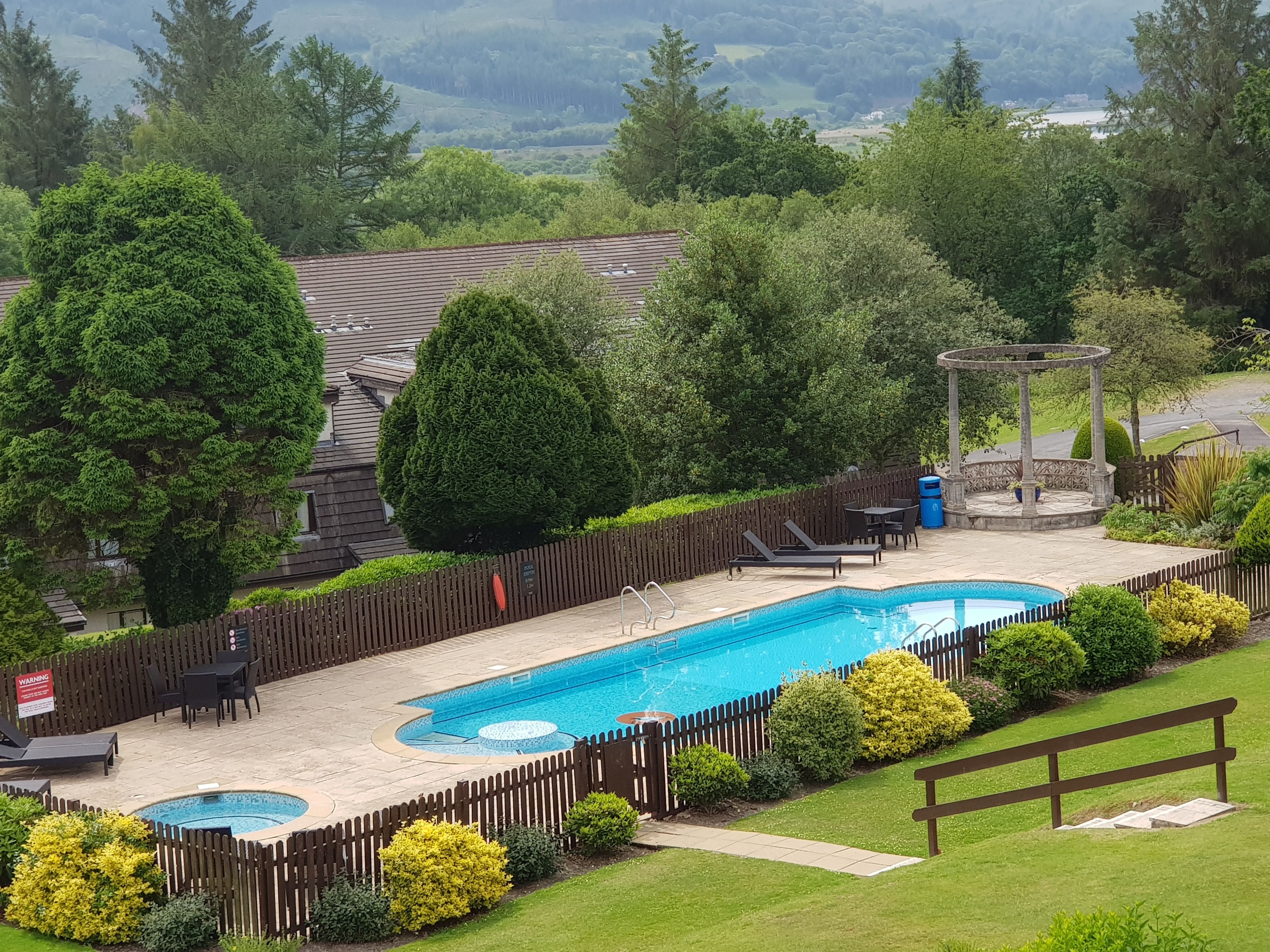 Une piscine rectangulaire avec une terrasse en bois, entourée d’une clôture en bois, et un kiosque en arrière-plan.