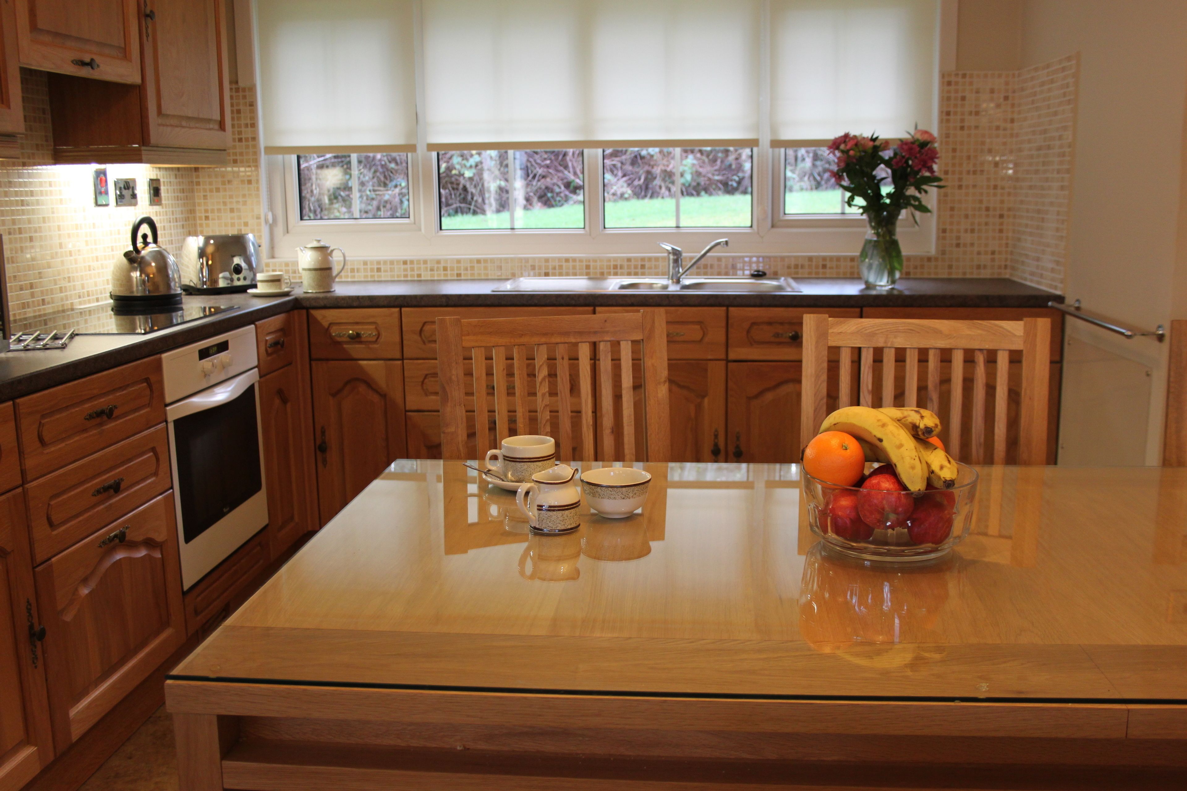 Une cuisine avec des meubles de rangement en bois, une table à plateau en verre et un panier de fruits.