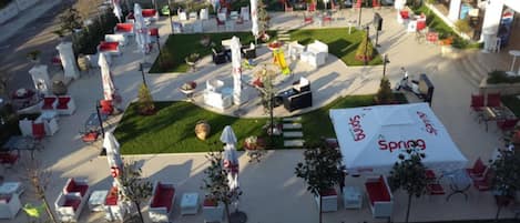 An outdoor seating area with white tables and red cushions, surrounded by white umbrellas and potted plants.