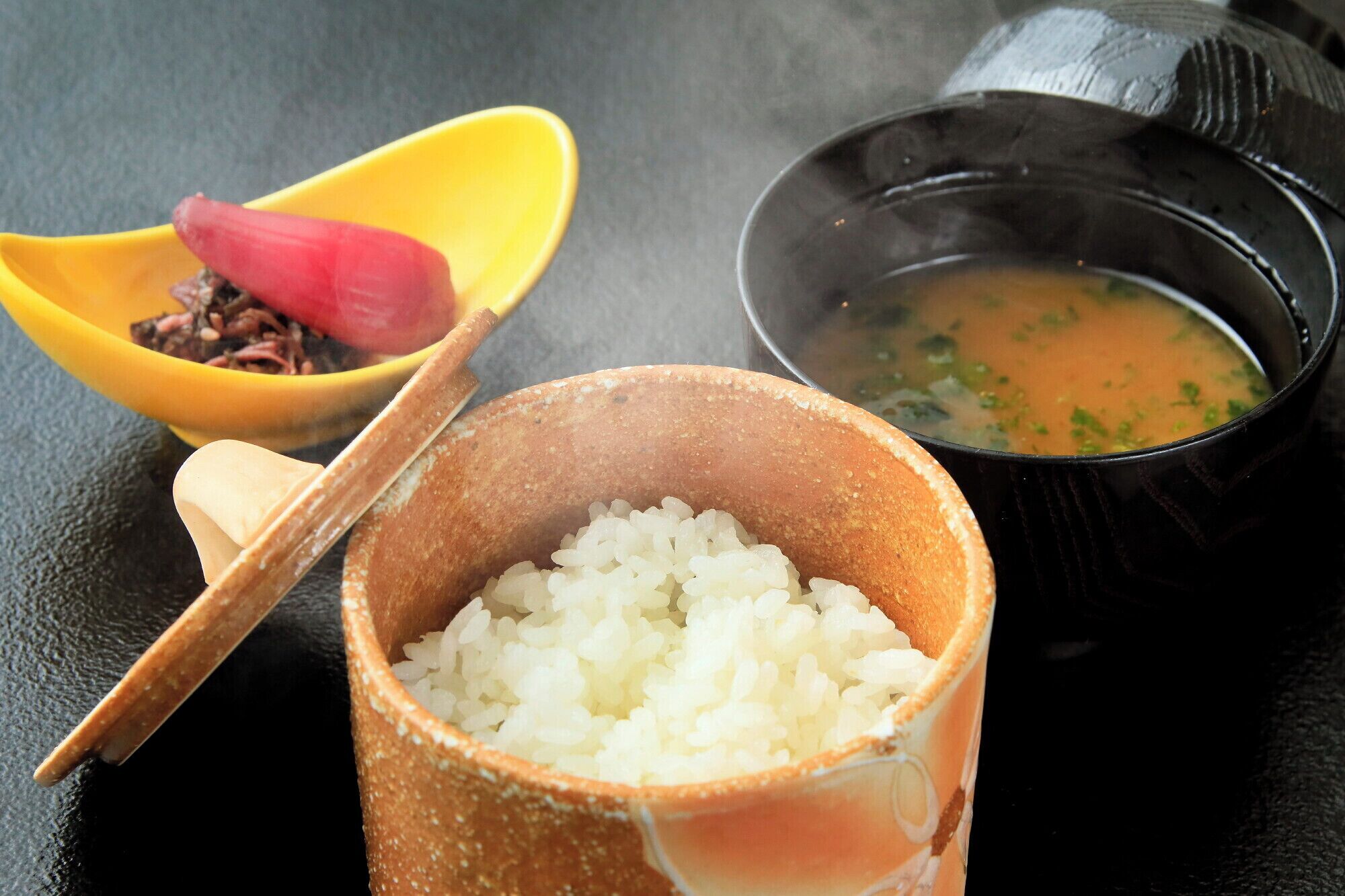 A bowl of white rice, a small bowl with a red object, and a black bowl with a liquid and green herbs.
