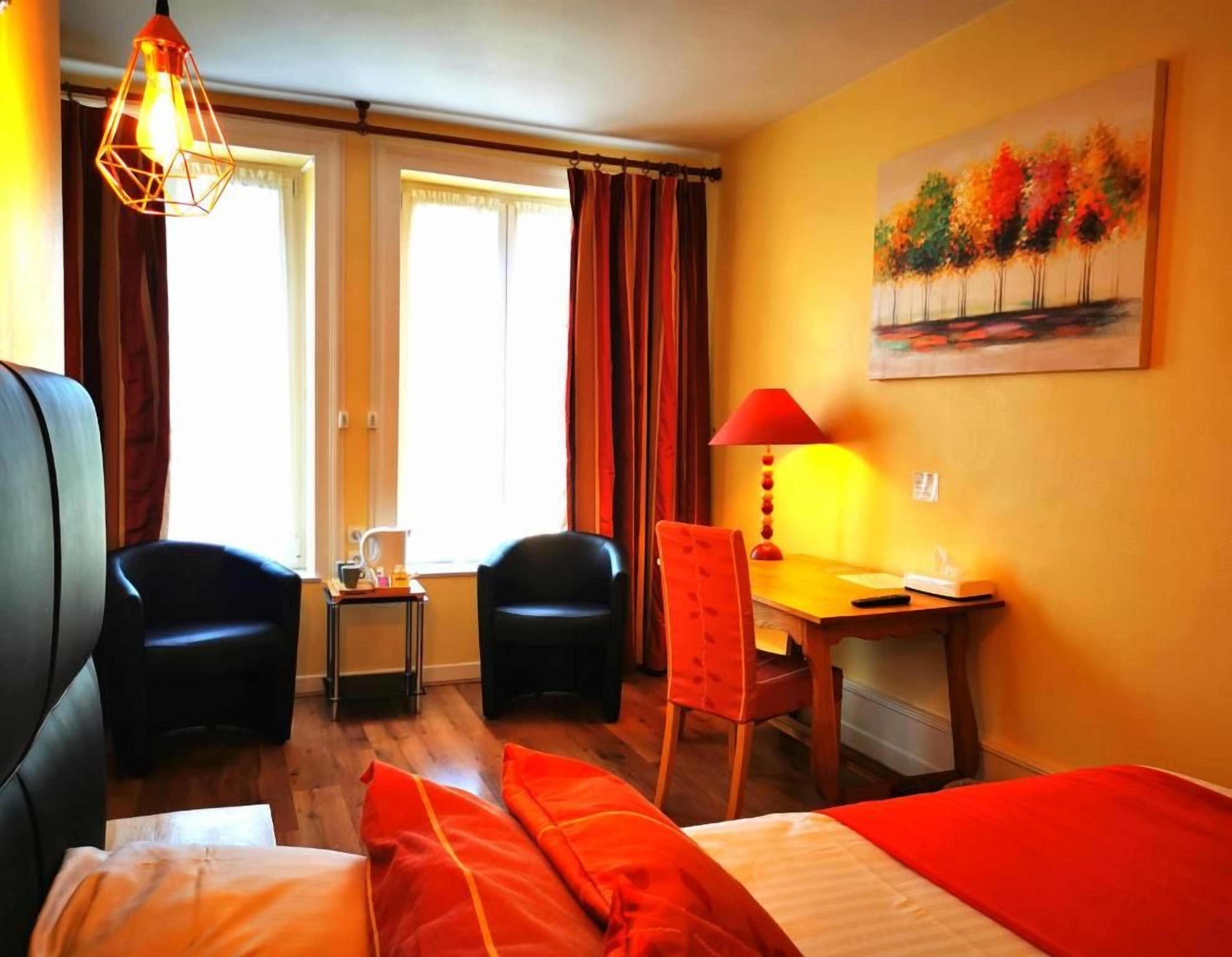 A hotel room with a bed, two chairs, a desk, and a painting on the wall.