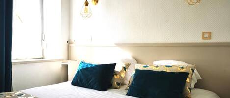A hotel room with a bed, two wall-mounted lamps, a window with curtains, and a bedside table.