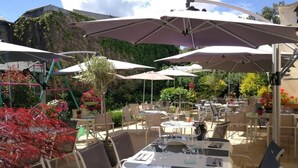 Een terras buiten met tafels, stoelen, parasols en potplanten.