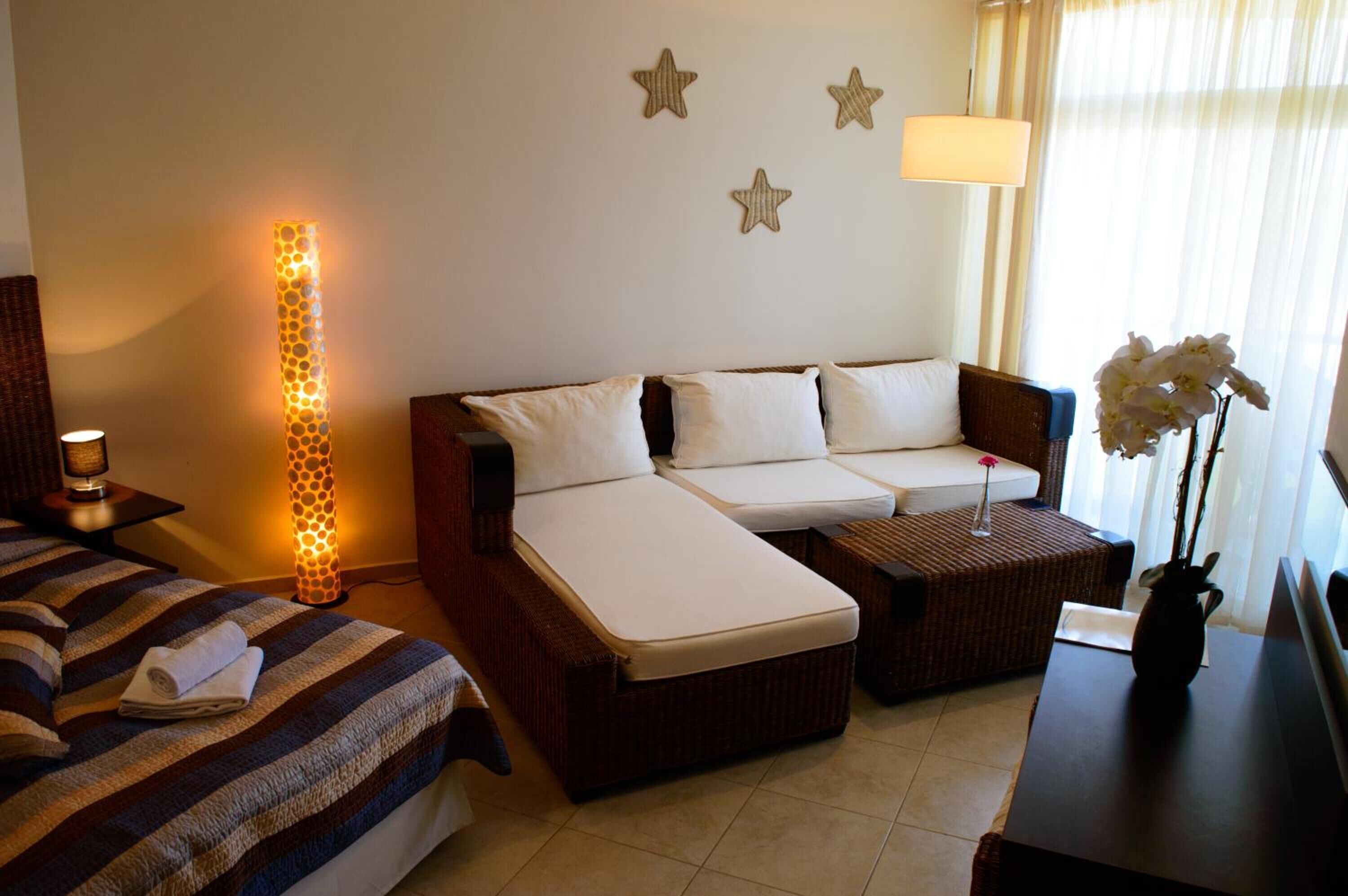 A hotel room with a bed, a sofa, and a small table. There is a lamp on the table and a vase with flowers on the table.