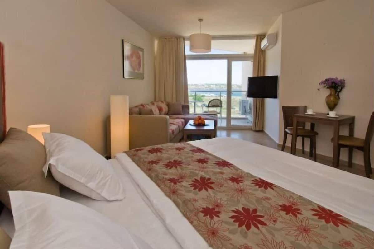 A hotel room with a bed, a sofa, a TV, a desk, and a balcony with a view.