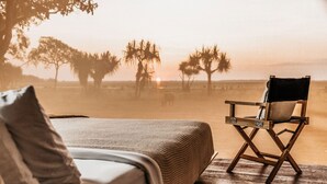 A bed with a blanket and pillows, a wooden deck, and a director's chair facing a sunset with palm trees in the background.