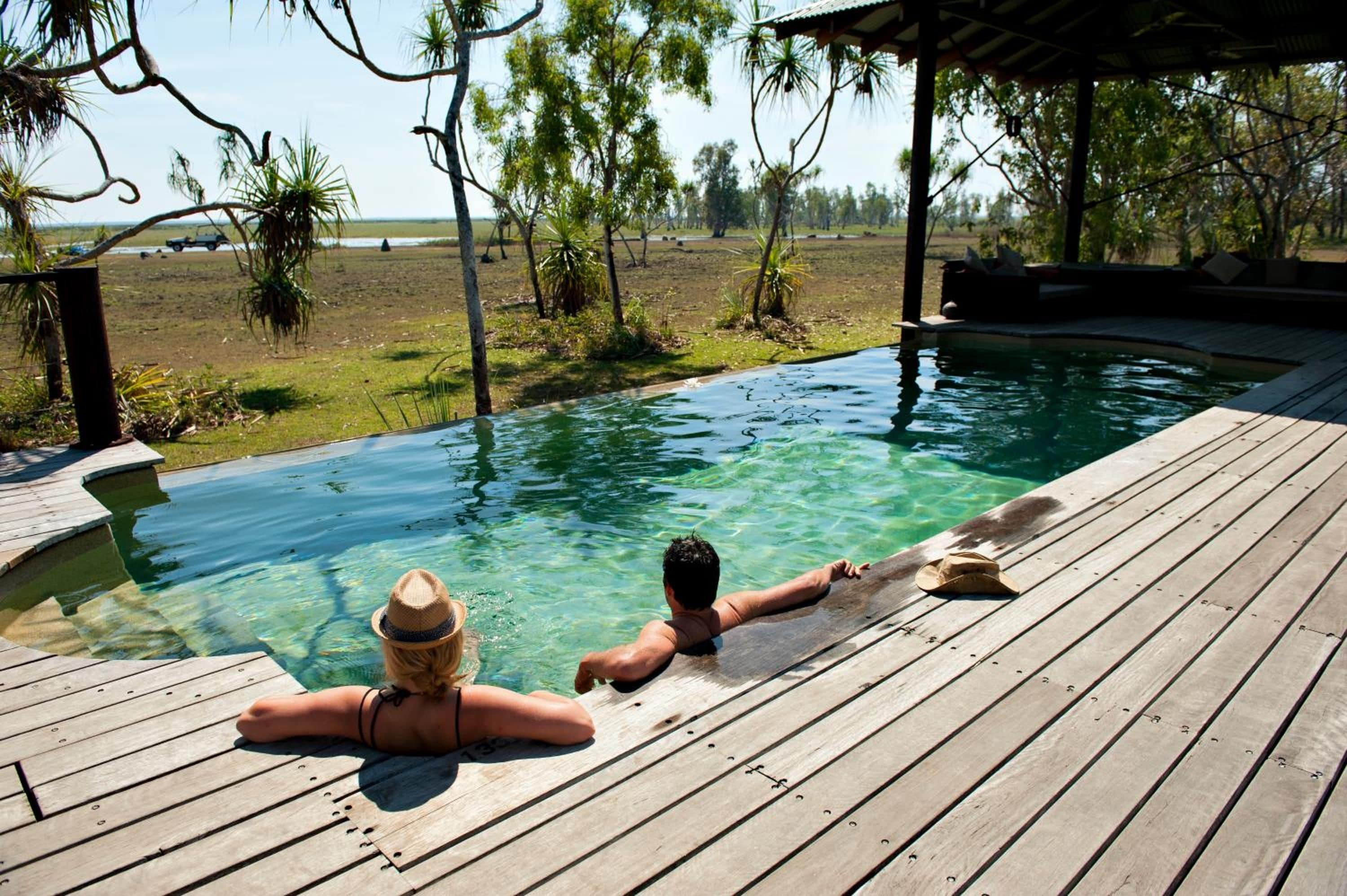 Une terrasse en bois avec une piscine à eau claire, entourée d’arbres et offrant une vue sur le paysage.
