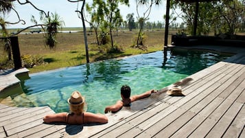 Une terrasse en bois avec une piscine à eau claire, entourée d’arbres et offrant une vue sur le paysage.