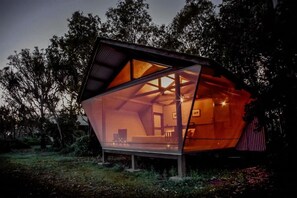 A modern, geometric glass structure with a visible interior, surrounded by trees.