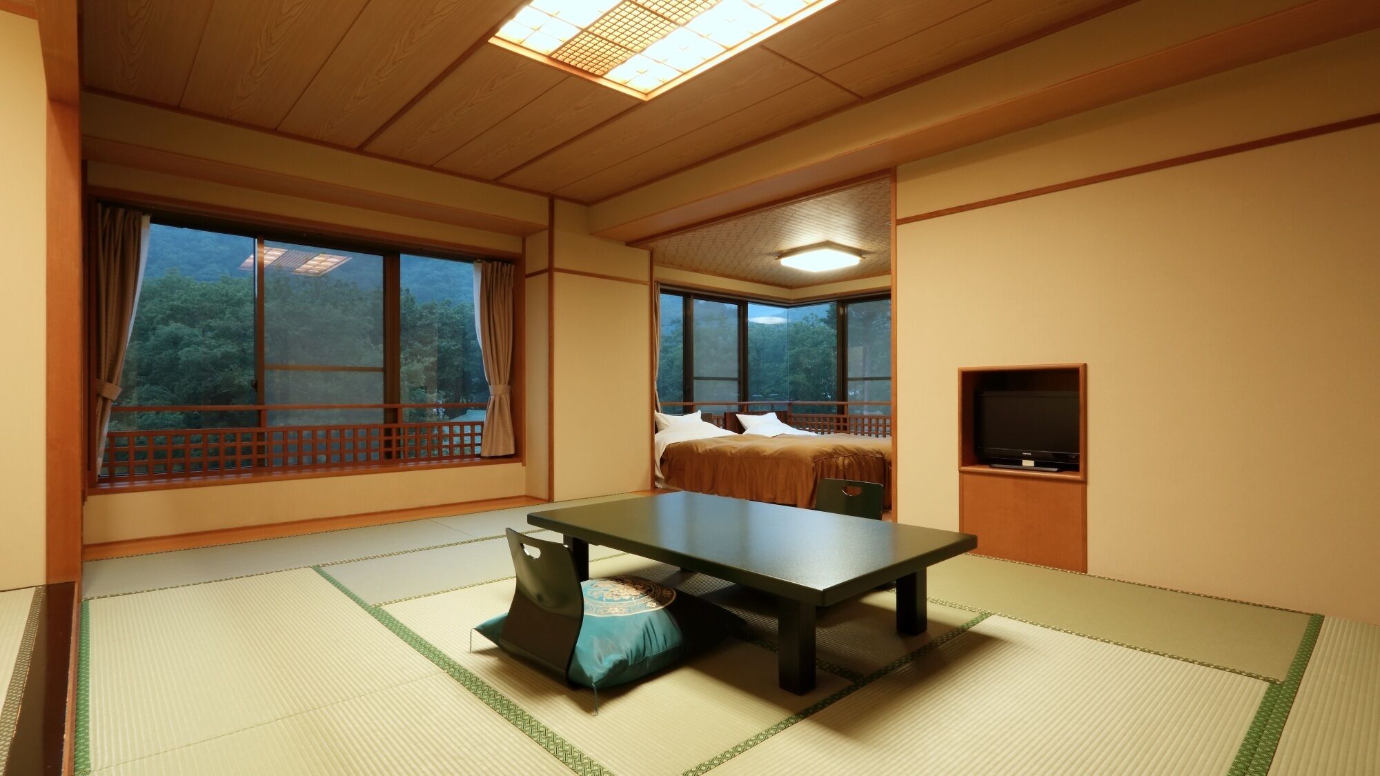 A room with tatami flooring, a bed with white bedding, a television, and large windows with a view of trees.