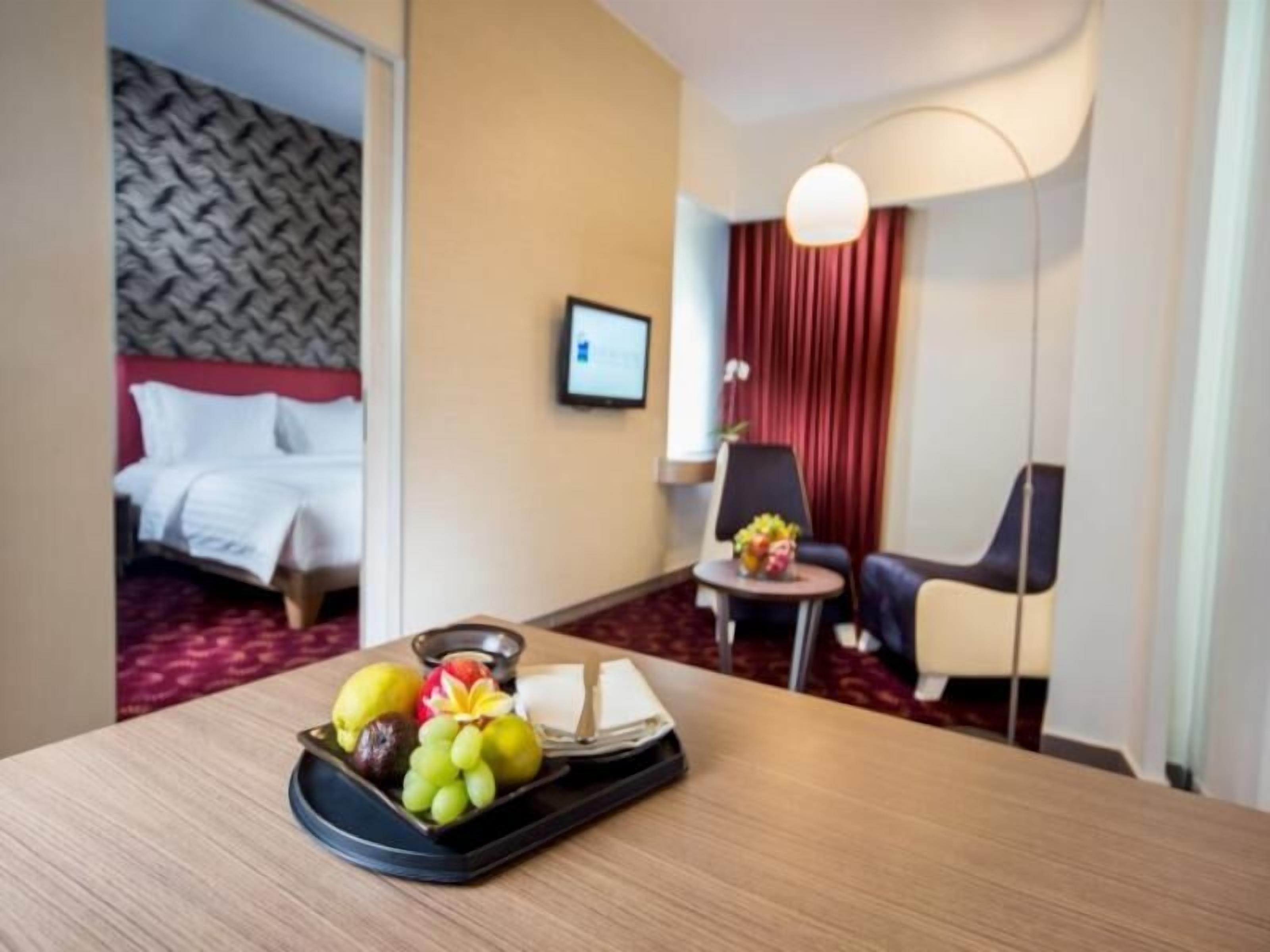 Une chambre d’hôtel avec un lit, un bureau sur lequel se trouvent des fruits et un plateau, une chaise et une lampe de bureau.