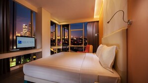 Quarto de hotel com uma cama grande, televisão em suporte, escrivaninha e vista para o horizonte noturno.