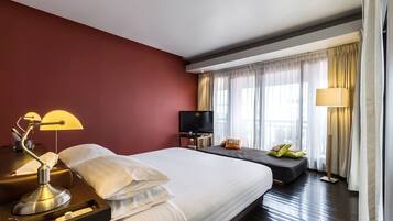Camera d'albergo con un letto grande, una scrivania con lampada, una televisione e un divano con cuscini.