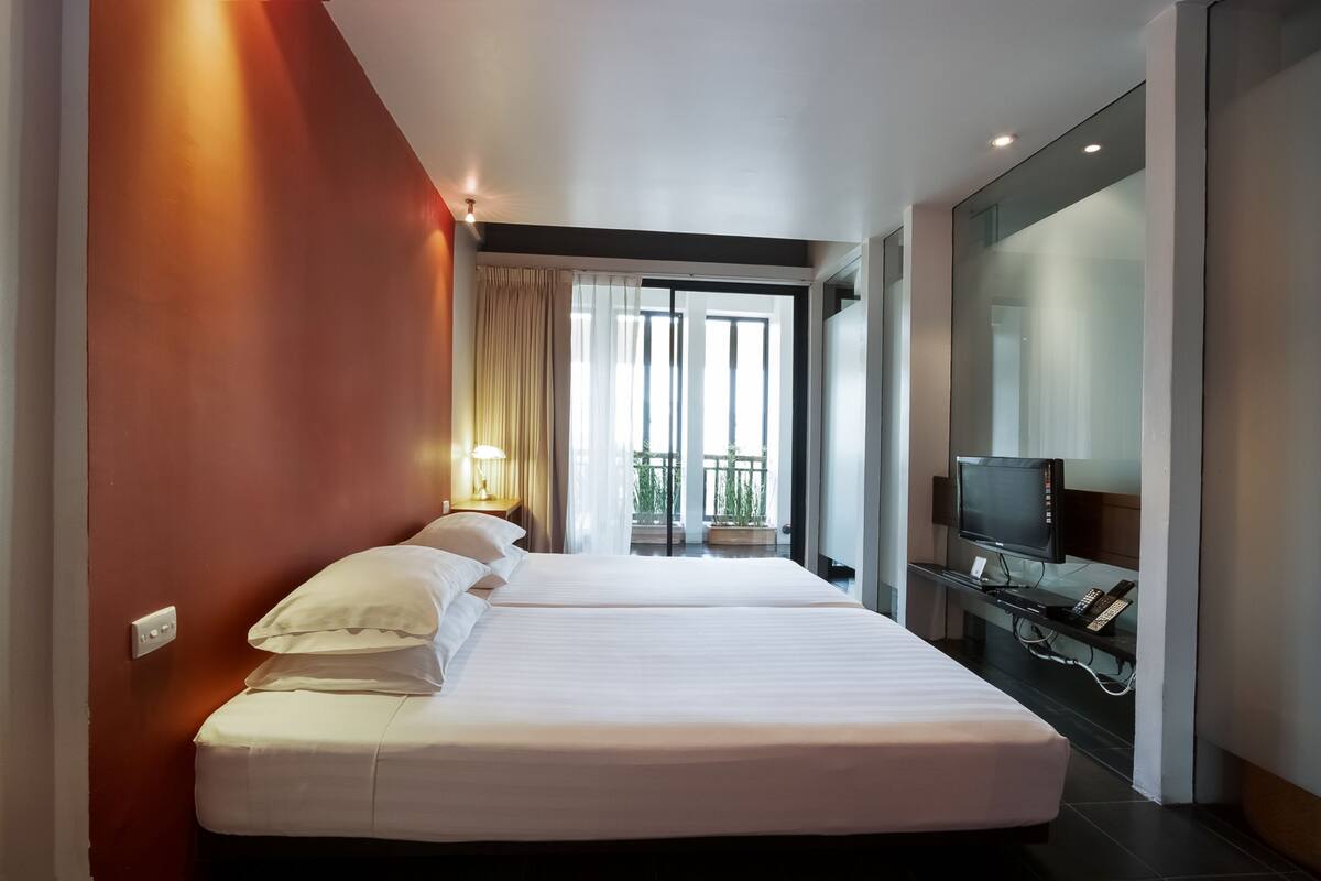 Habitación de hotel con dos camas, televisor, escritorio con computadora y un amplio ventanal con vista al exterior.
