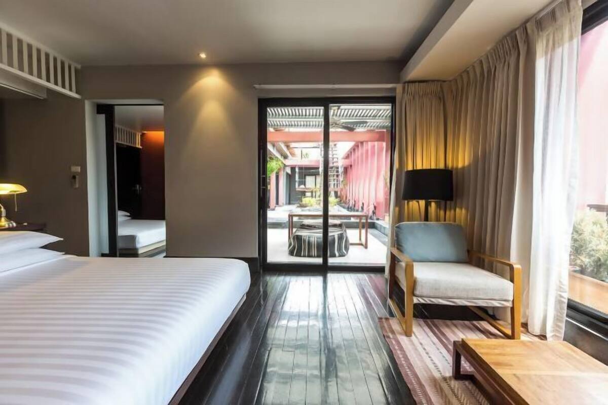 Habitación de hotel con cama, una silla, una mesita pequeña y un ventanal corredero que da a un espacio exterior con mesa y sillas.
