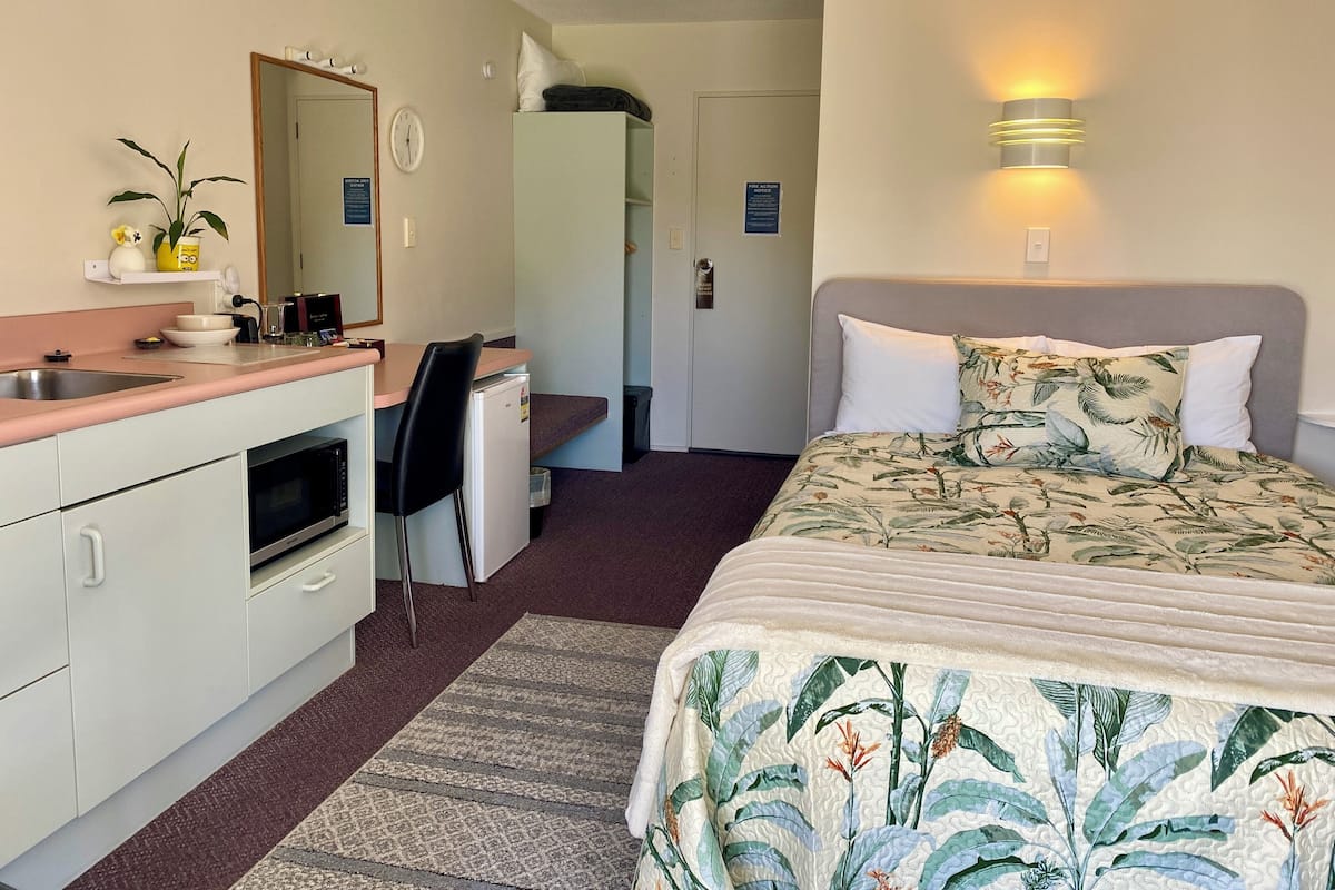 A compact hotel room with a kitchenette, a bed with floral bedding, a desk with a chair, a mirror, and a wall-mounted light fixture.