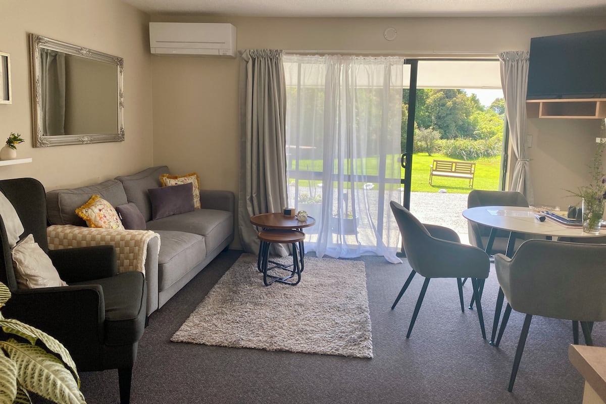 A living room with a grey sofa, a small round table, and a dining area with chairs. There is a large window with curtains, an air conditioning unit on the wall, and a mirror above the sofa.