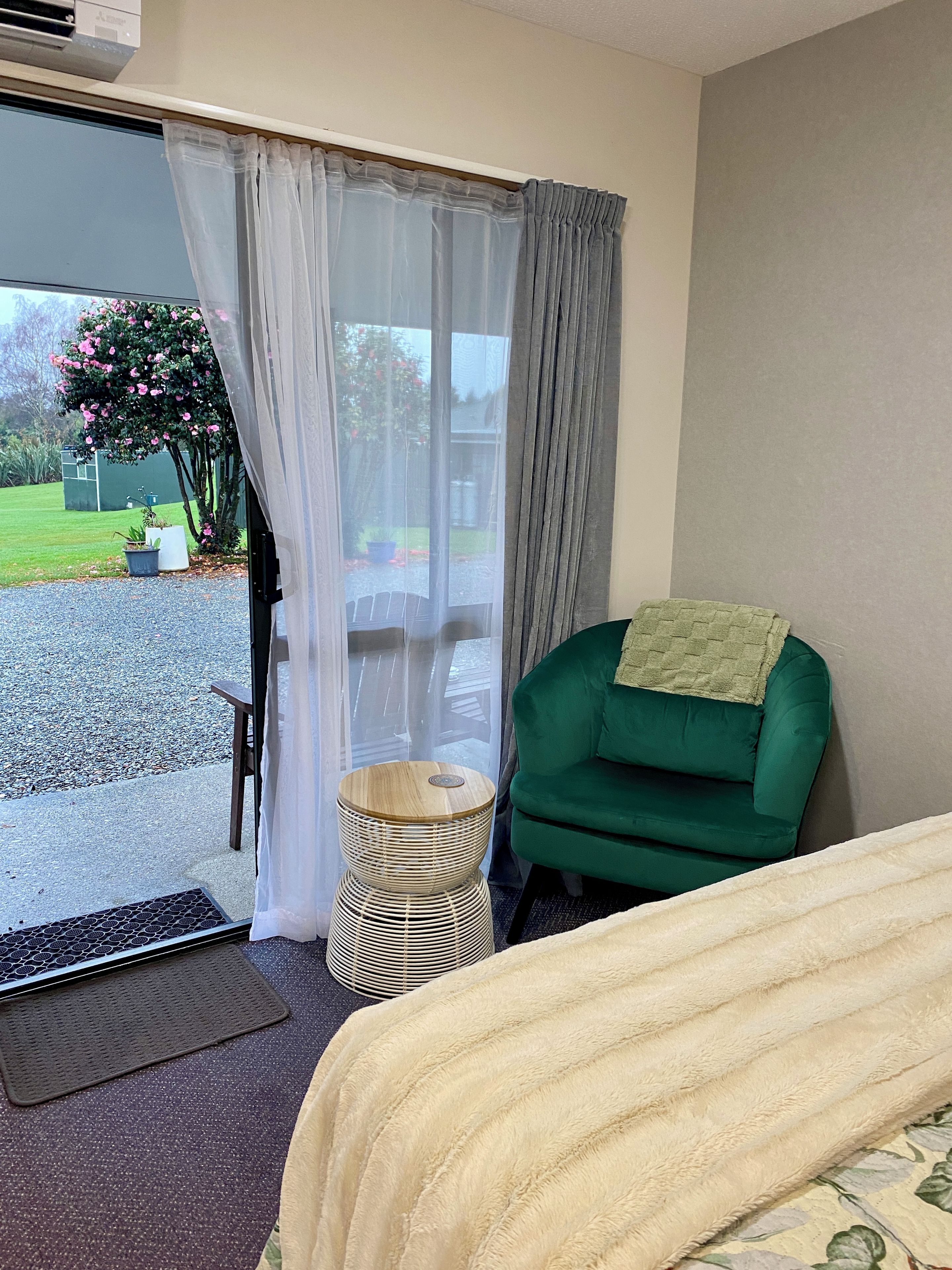 A hotel room with a green armchair, a small round table, a bed with a white cover, and a view of a garden through a sliding door.
