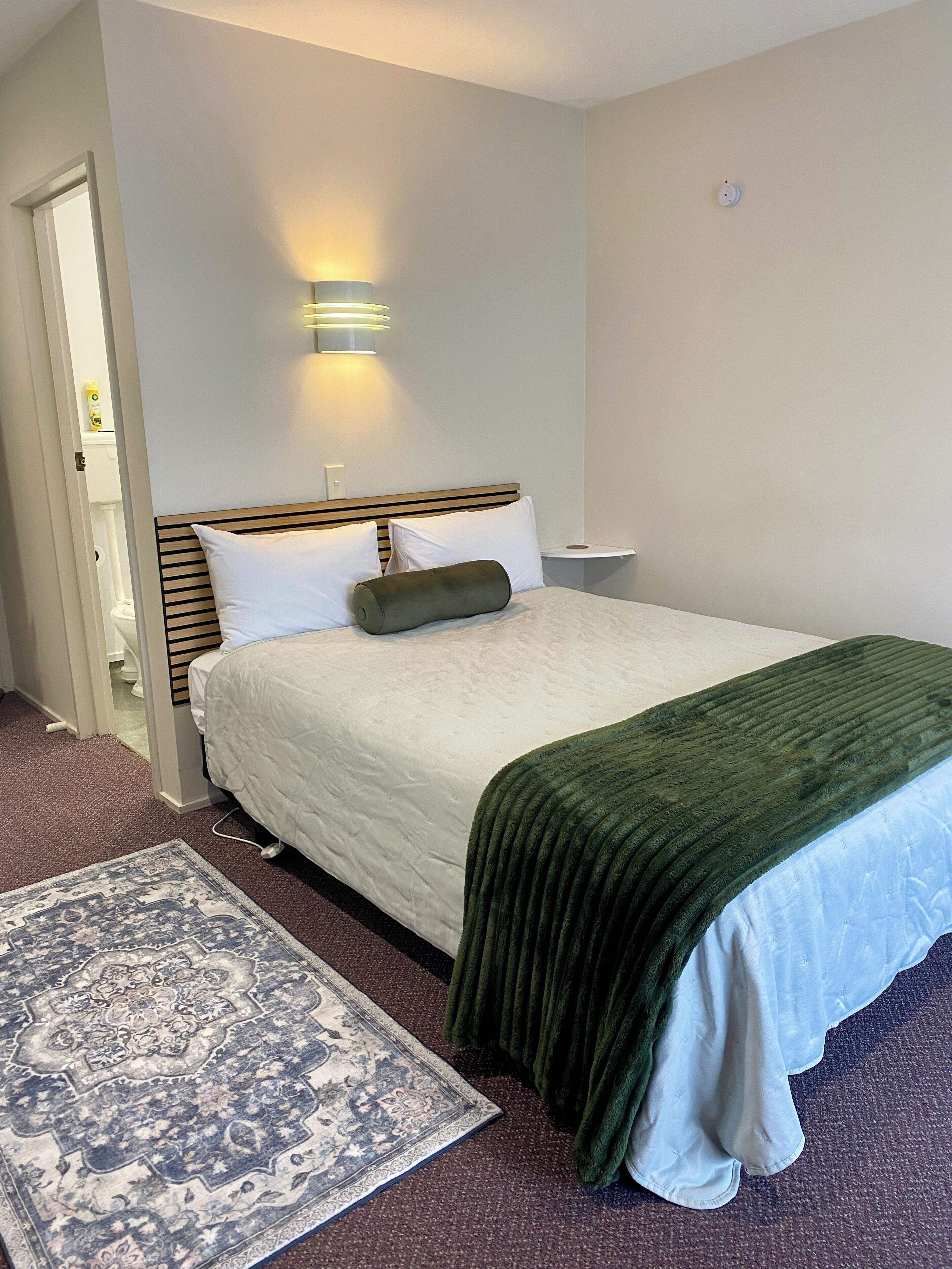 Habitación de hotel con cama, mesita de noche, lámpara de pared y alfombra estampada.