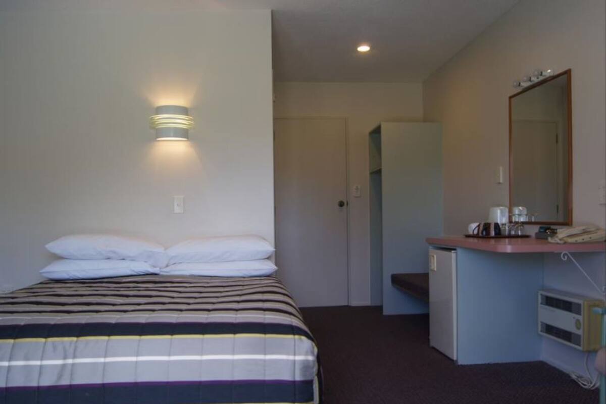 A hotel room with a bed, a bedside lamp, a small desk with a mirror, and a door leading to another room.