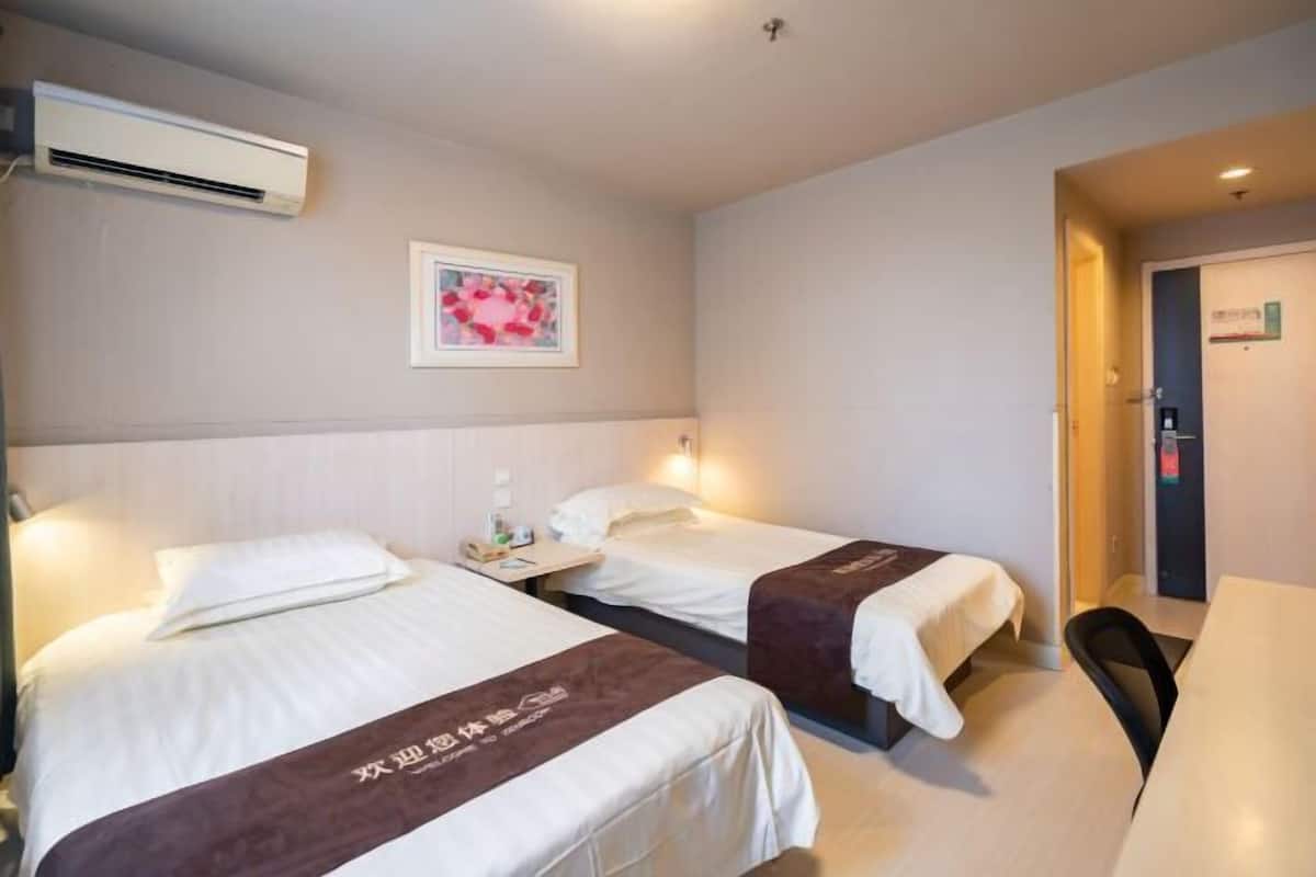 A hotel room with two beds, a desk, and a chair. There is a wall-mounted air conditioner and a framed picture above the beds.