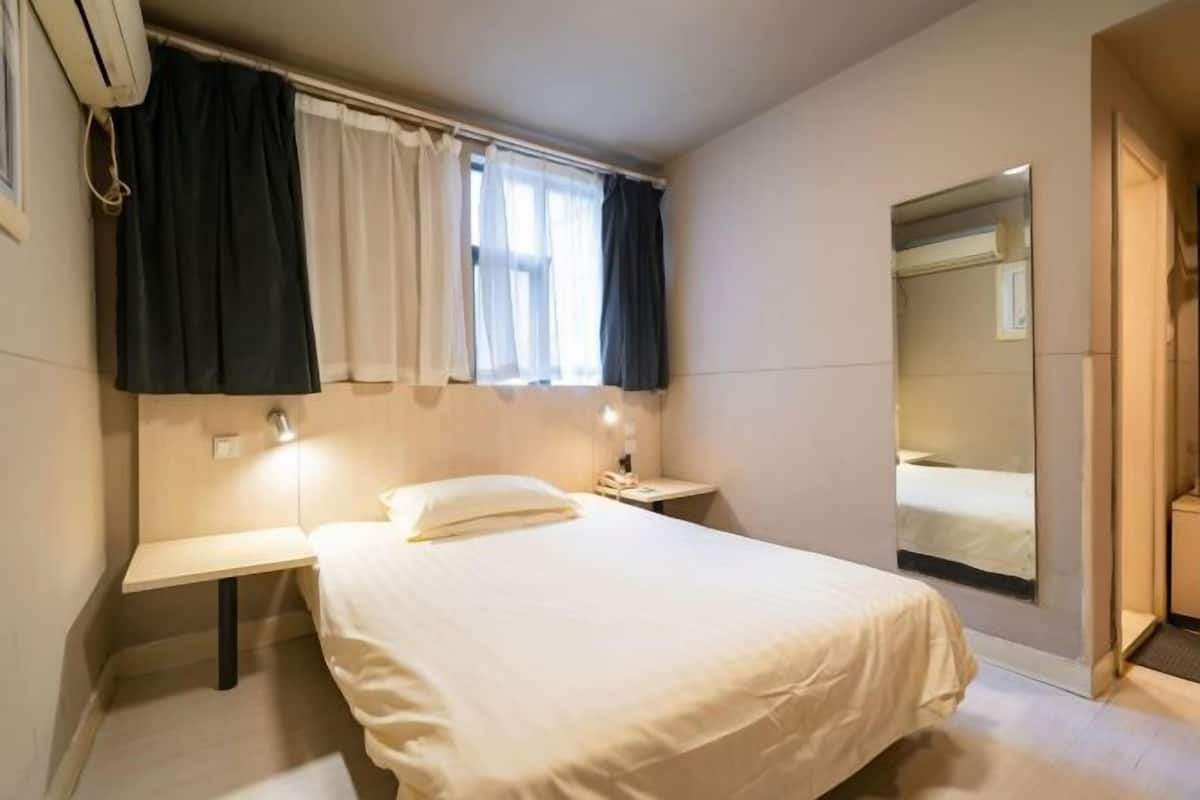 A bedroom with a bed, bedside table, lamp, window with curtains, and a mirror.