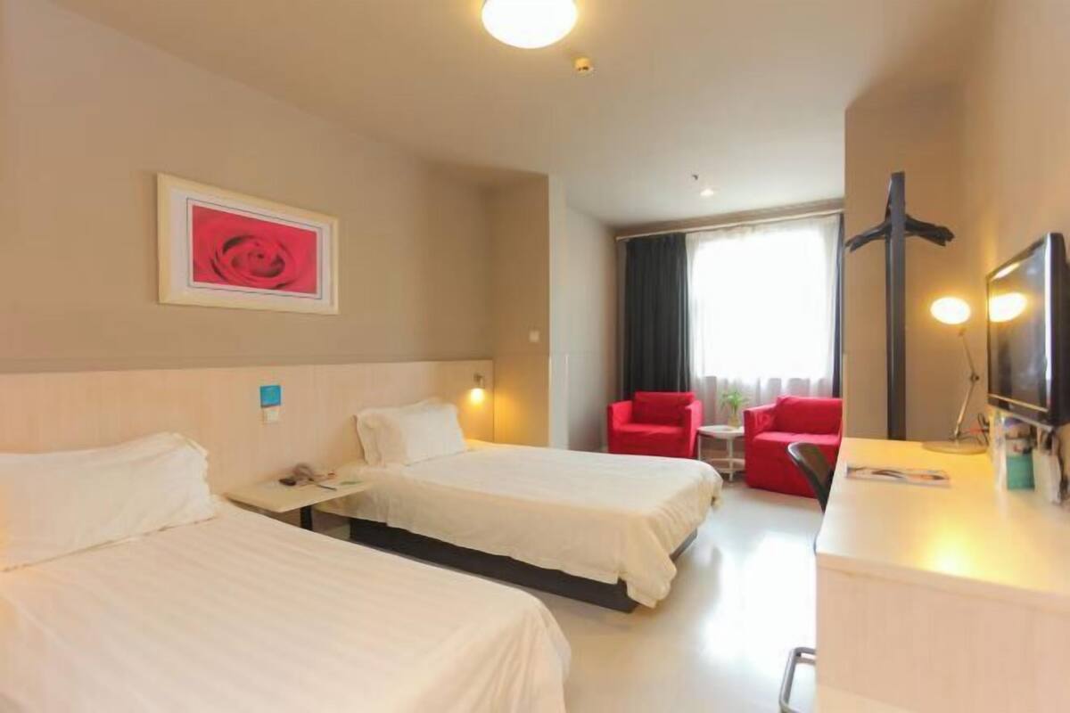A hotel room with two beds, a desk, a red chair, a TV, and a large window.