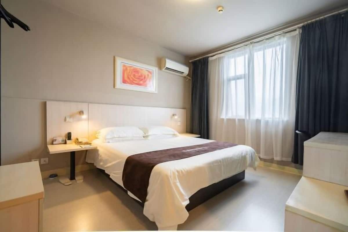 A hotel room with a bed, bedside table, lamp, air conditioning unit, and a window with curtains.