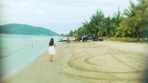 A beach with a person in a white dress walking away from the camera, palm trees, and a vehicle parked on the sand. - Laemsing Whitehouse Resort (Laem Sing)