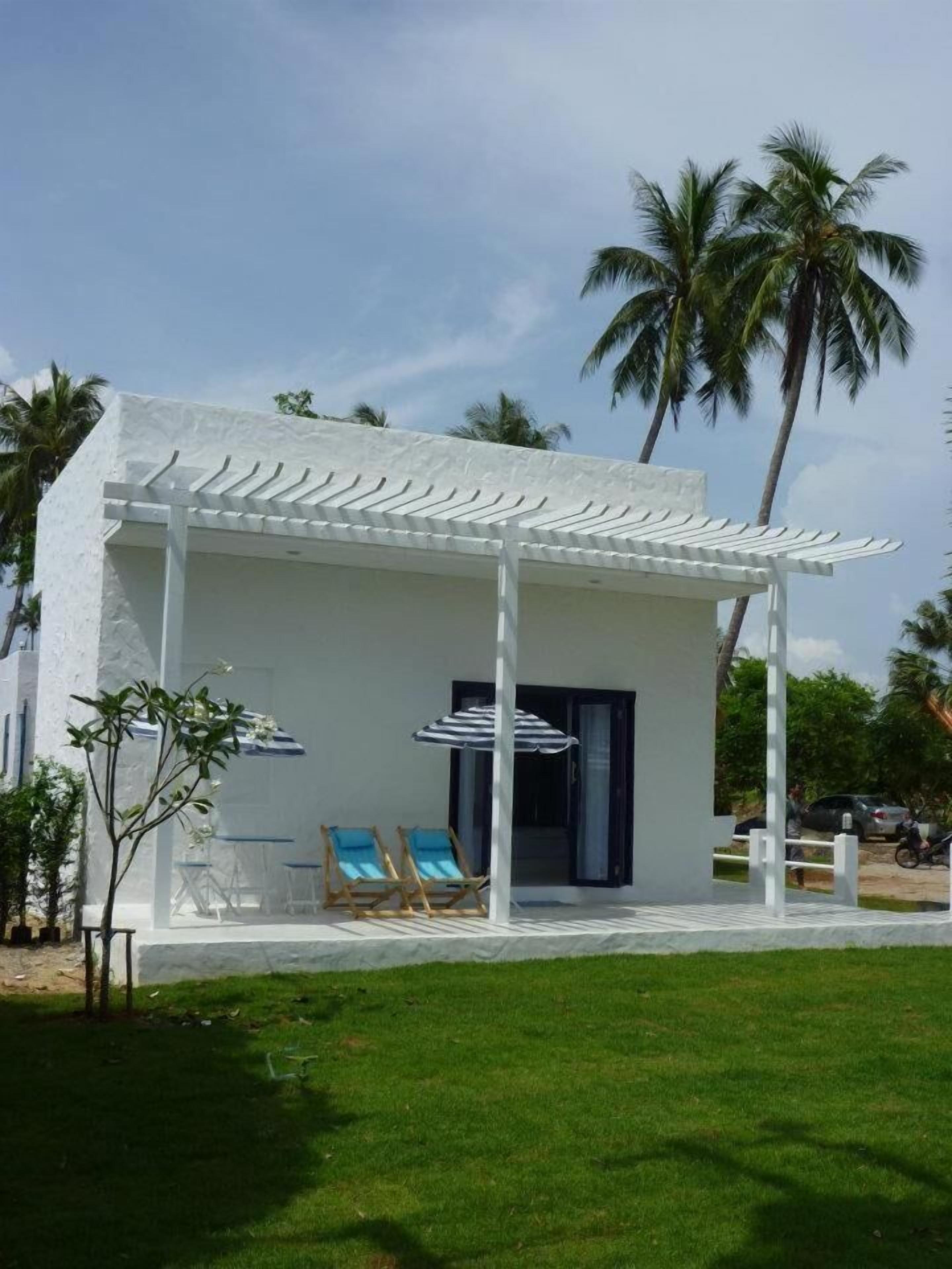 Un edificio blanco con un porche cubierto, dos sillones de descanso y una sombrilla a rayas.