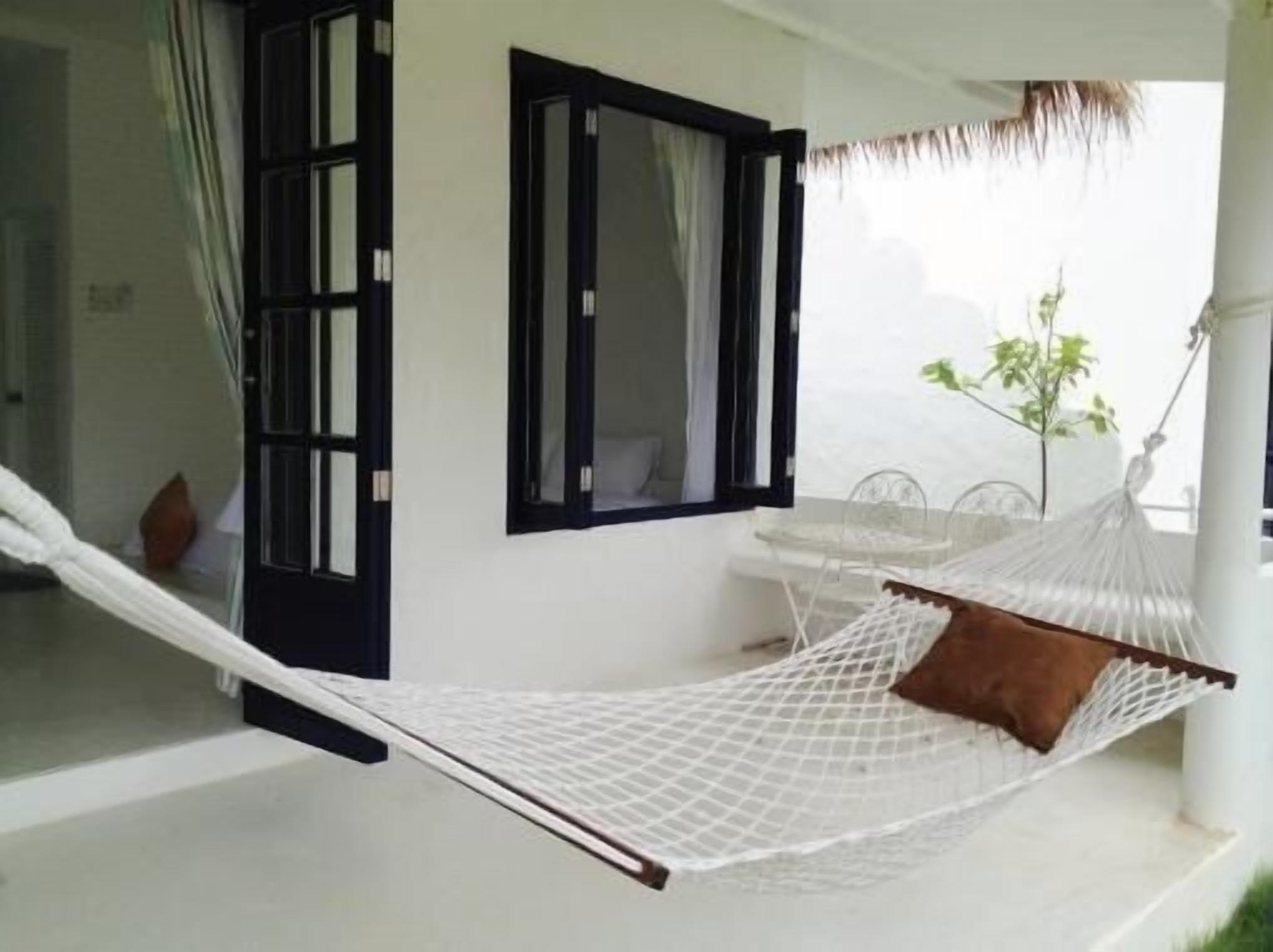 A hammock on a balcony with a white railing, a window with black shutters, and a small table with a chair.