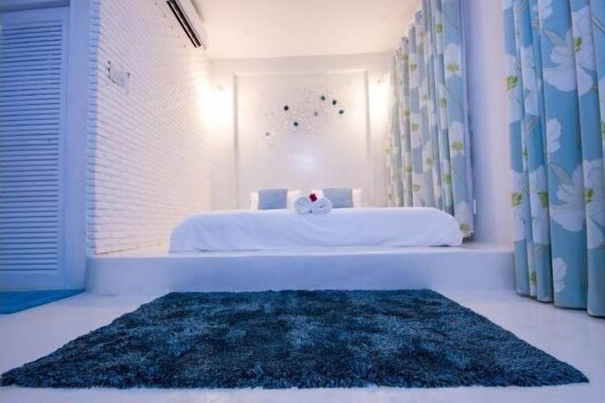 Un dormitorio con cama, alfombra azul estampada, paredes blancas y ventana con cortinas.