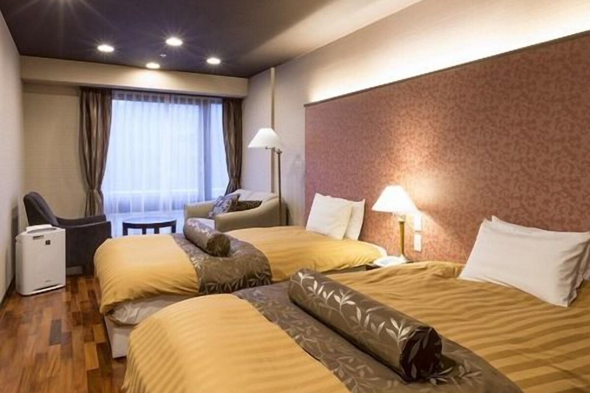 Ein Hotelzimmer mit zwei Betten, einem Schreibtisch, einem Stuhl, einer Lampe und einem Fenster mit Vorhängen.
