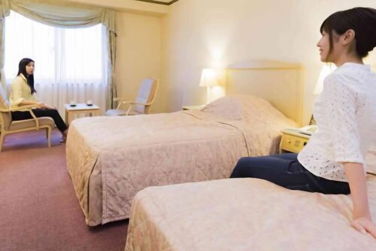 Ein Hotelzimmer mit zwei Betten, einem Sessel, einem Nachttisch und einem Fenster mit Vorhängen.